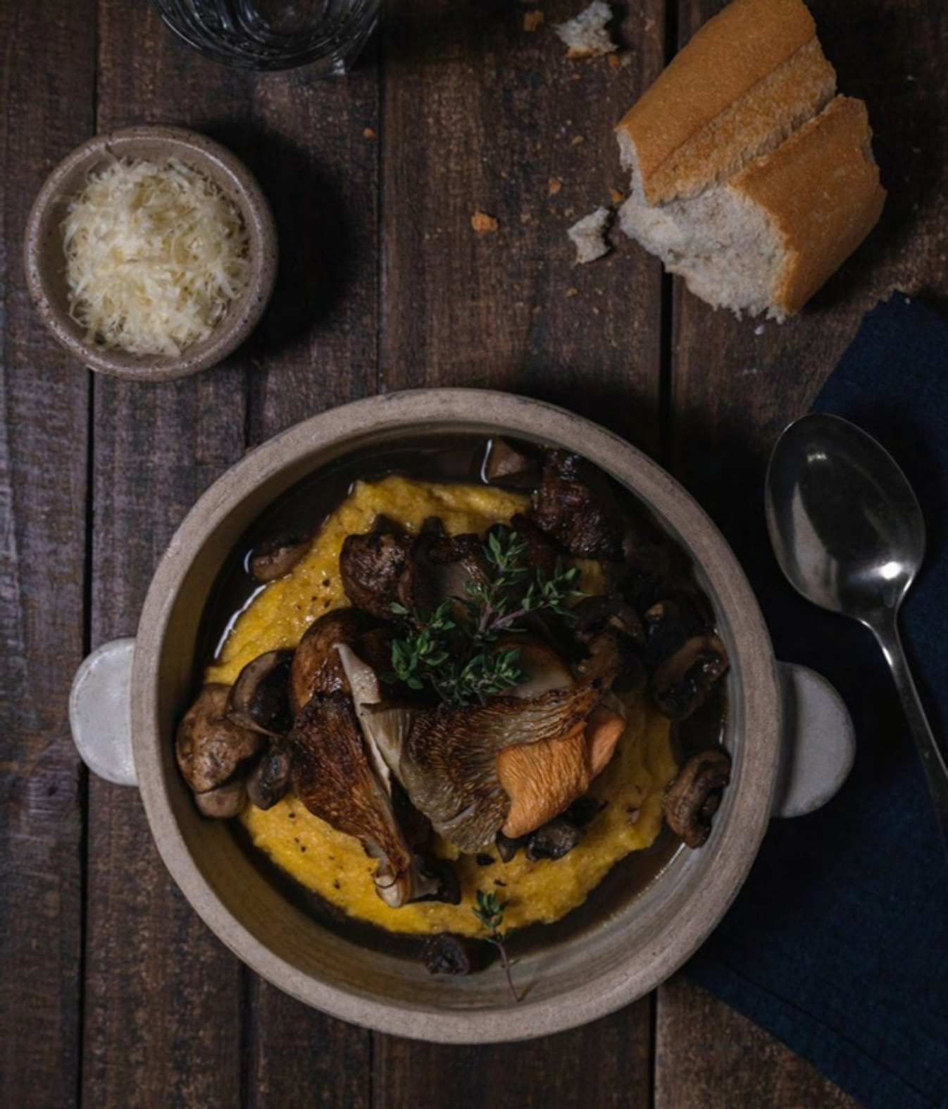 Cazuela de estofado de hongos con polenta, pan y queso rallado sobre mesa de madera