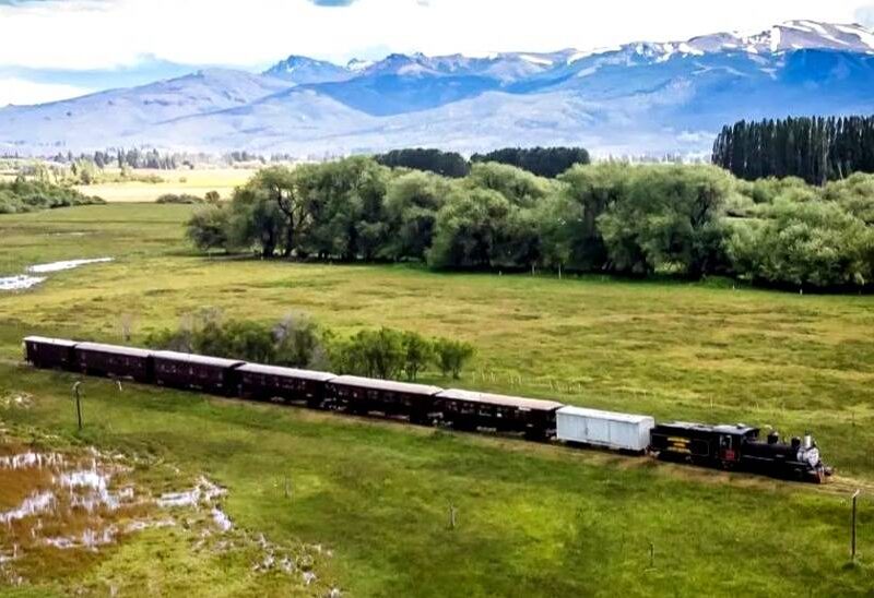 Vista aérea de La Trochita, el Viejo Expreso Patagónico, cruzando un valle verde con la cordillera nevada de fondo en Chubut
