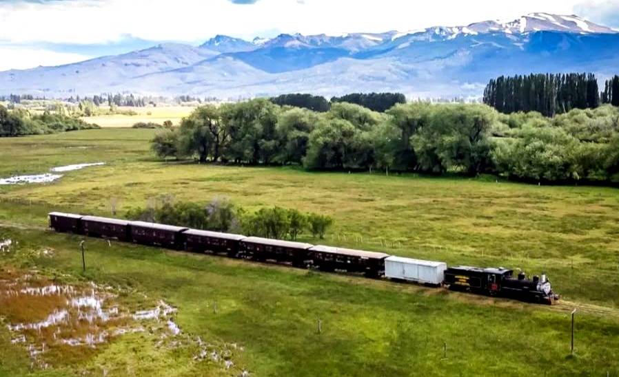 Vista aérea de La Trochita, el Viejo Expreso Patagónico, cruzando un valle verde con la cordillera nevada de fondo en Chubut