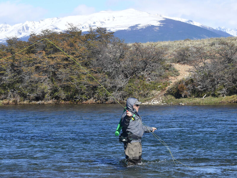Pescador lanzando con caña de mosca en un río de la cordillera de Chubut, con montañas nevadas de fondo