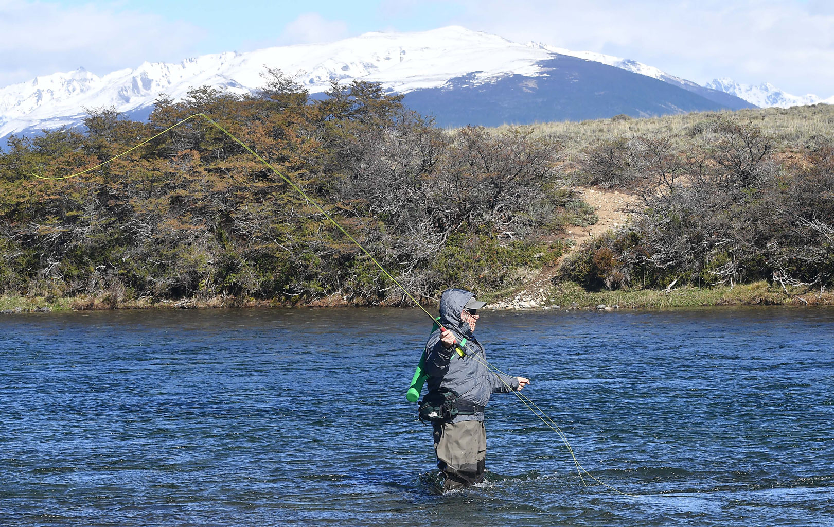 Pescador lanzando con caña de mosca en un río de la cordillera de Chubut, con montañas nevadas de fondo
