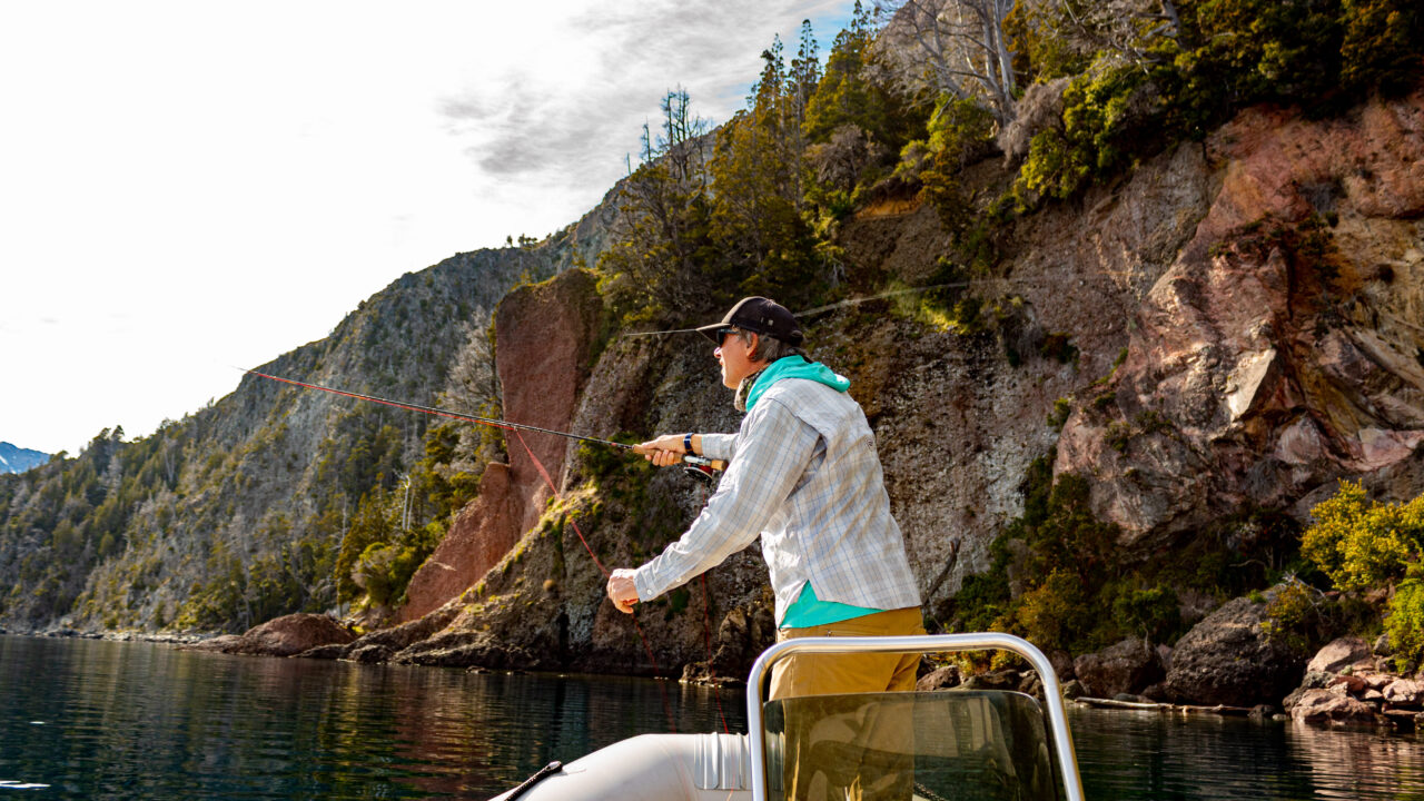 Pescador con caña de mosca lanzando desde un bote en el lago Nahuel Huapi, frente a acantilados con bosque en Villa La Angostura