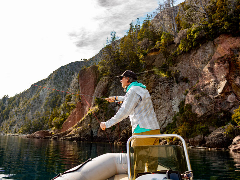Pescador con caña de mosca lanzando desde un bote en el lago Nahuel Huapi, frente a acantilados con bosque en Villa La Angostura