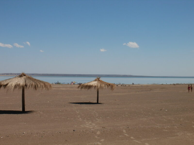 Playa de arena con sombrillas de paja a orillas del lago del embalse Casa de Piedra, en el sudoeste de La Pampa