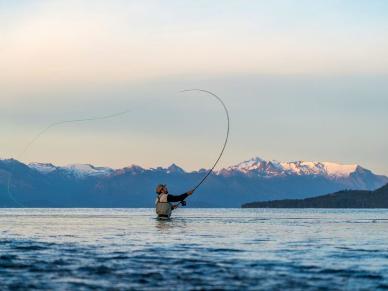 Pescador con mosca lanzando la línea dentro del lago Nahuel Huapi, con la cordillera nevada al atardecer
