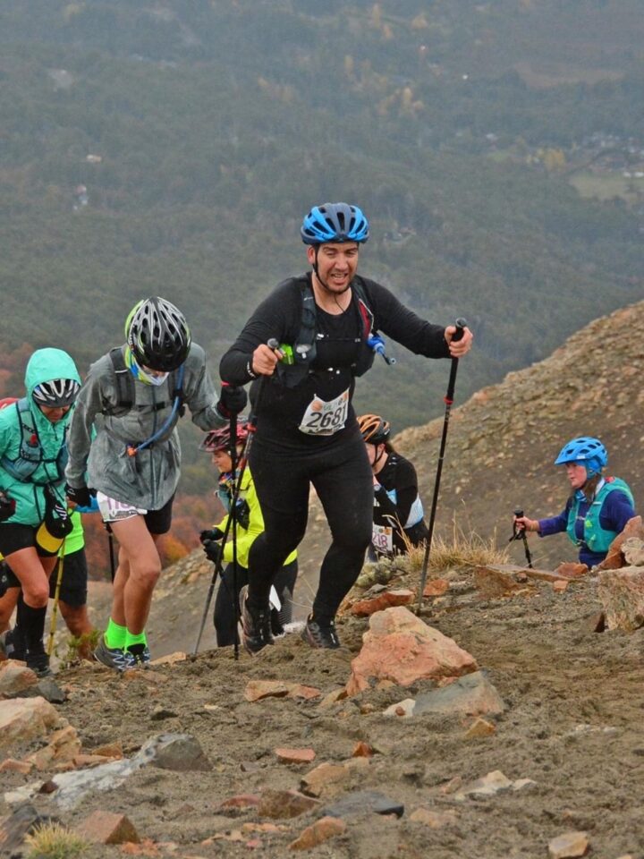 Corredores con bastones suben una ladera de piedra durante la Travesía de los Cerros en Villa La Angostura, con el bosque y el pueblo de fondo